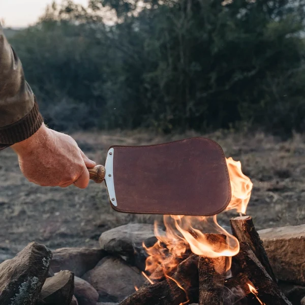 Perfect Hideaways: Donkey Fire Paddle. A hand holding a leather and wood fire blower paddle over a burning campfire surrounded by rocks, set against a backdrop of wild bushveld. Perfect Hideaways shop
