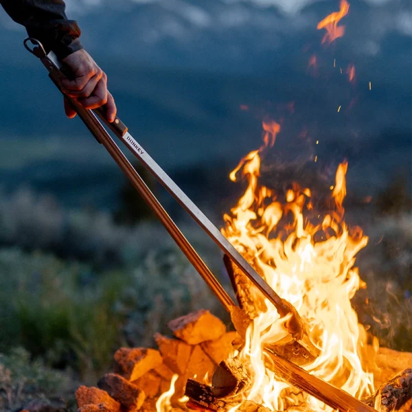 Perfect Hideaways: Donkey long tongs in use at a campfire, turning logs in the flames with mountains in the background. Perfect Hideaways shop