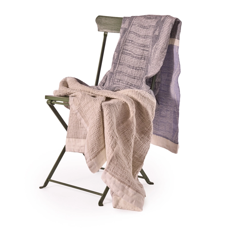 Perfect Hideaways: A Ocean Bogolan Lounge Throw in soft beige and lavender draped over a rustic green metal chair, showcasing its elegant woven pattern and gentle folds. Perfect Hideaways shop