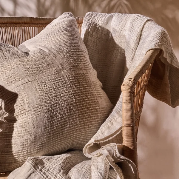 Perfect Hideaways: Textured natural sand-coloured Bogolan Continental Cushion Cover and throw styled on a woven rattan chair in warm, dappled lighting. Perfect Hideaways shop