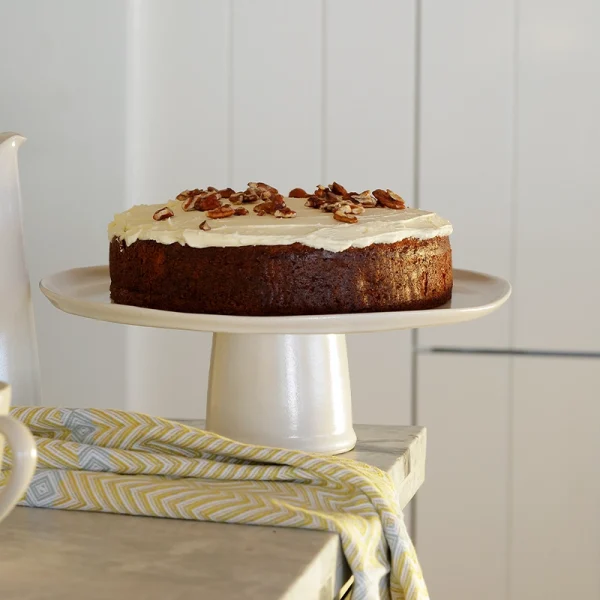 Perfect Hideaways: A freshly frosted carrot cake topped with pecans, displayed on a ceramic pedestal cake stand in a sunlit kitchen. The shop