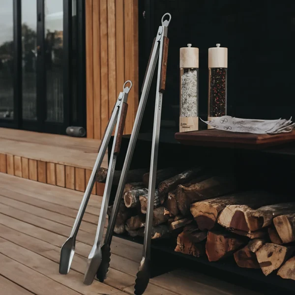 Perfect Hideaways: Two Donkey Tong braai tongs resting against a deck-side counter with wood storage and spice grinders, styled for a rustic outdoor setting. Perfect Hideaways shop