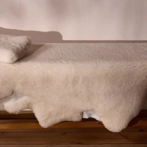 Perfect Hideaways: A plush, cream-coloured sheepskin throw draped over a wooden bench with a matching folded sheepskin cushion, set against a soft pink-toned wall with natural shadows. Perfect Hideaways shop