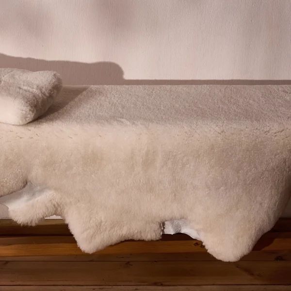 Perfect Hideaways: A plush, cream-coloured sheepskin throw draped over a wooden bench with a matching folded sheepskin cushion, set against a soft pink-toned wall with natural shadows. Perfect Hideaways shop