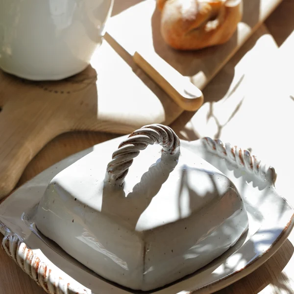 Perfect Hideaways: Sunlight casts dappled shadows over a ceramic butter dish with hand-twisted handles, surrounded by fresh bread and a wooden board – Perfect Hideaways shop