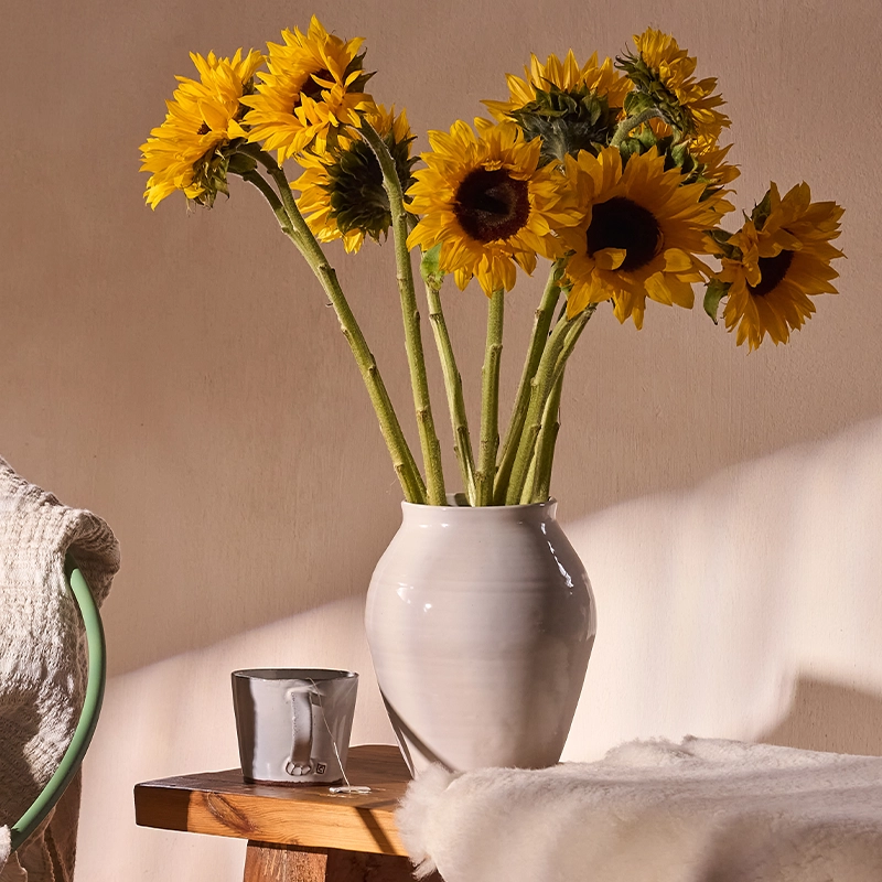 Perfect Hideaways: A white ceramic vase filled with vibrant yellow sunflowers sits atop a rustic wooden side table, beside a grey mug and a textured throw, basking in soft natural light. Perfect Hideaways shop