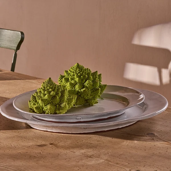 Perfect Hideaways: A rustic wooden table set with a trio of handcrafted oval ceramic platters in neutral tones, elegantly styled with Romanesco broccoli for a natural touch – bathed in warm afternoon light with shadows on a plastered wall. Perfect Hideaways shop.