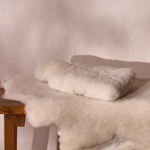 Perfect Hideaways: A neatly folded sheepskin hot water bottle cover rests on a matching sheepskin throw atop a wooden bench, showcasing its luxurious texture in a serene, neutral-toned setting. Perfect Hideaways shop