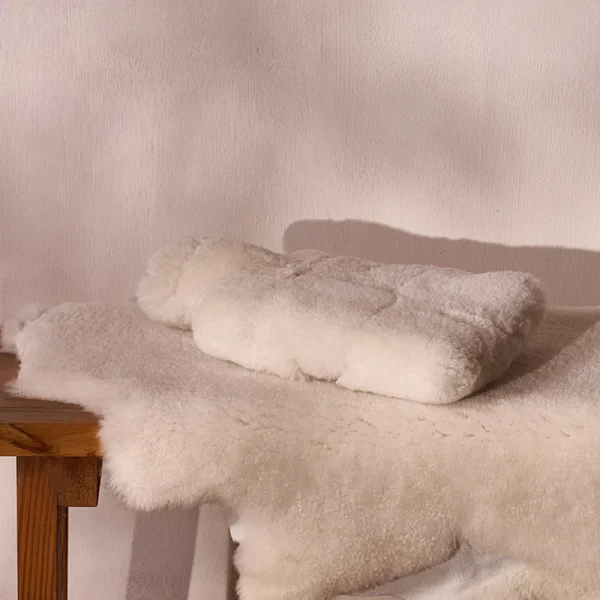 Perfect Hideaways: A neatly folded sheepskin hot water bottle cover rests on a matching sheepskin throw atop a wooden bench, showcasing its luxurious texture in a serene, neutral-toned setting. Perfect Hideaways shop
