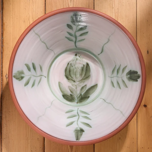 Perfect Hideaways: Top-down view of a hand-painted terracotta bowl with a large central protea motif surrounded by leafy stems in green tones against a pale interior glaze. Perfect Hideaways shop.
