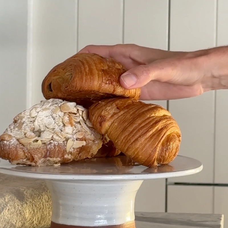 Perfect Hideaways: A tempting trio of buttery croissants and an almond pastry rest on a ceramic cake stand, as a hand reaches in to pick one — a perfect blend of rustic indulgence and artisanal charm. Perfect Hideaways shop.