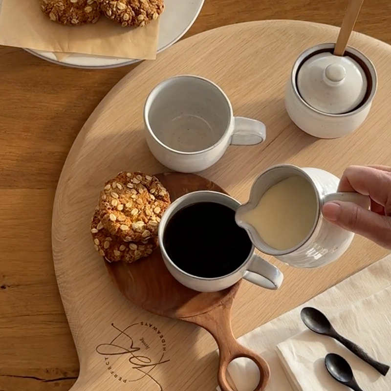 Perfect Hideaways: A warm, inviting coffee moment featuring rustic stoneware mugs, homemade oat cookies, and cream being poured into black coffee on a beautifully crafted wooden board — a scene of slow living and morning indulgence. Perfect Hideaways shop.
